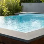 Vue rapprochée d'un spa moderne avec de l'eau bleue claire, bordé de bois et de pierre grise, sur fond de verdure luxuriante.