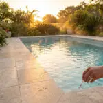 Main tenant une bandelette de test dans l'eau d'une piscine bordée de dalles claires et de végétation luxuriante, au coucher du soleil.